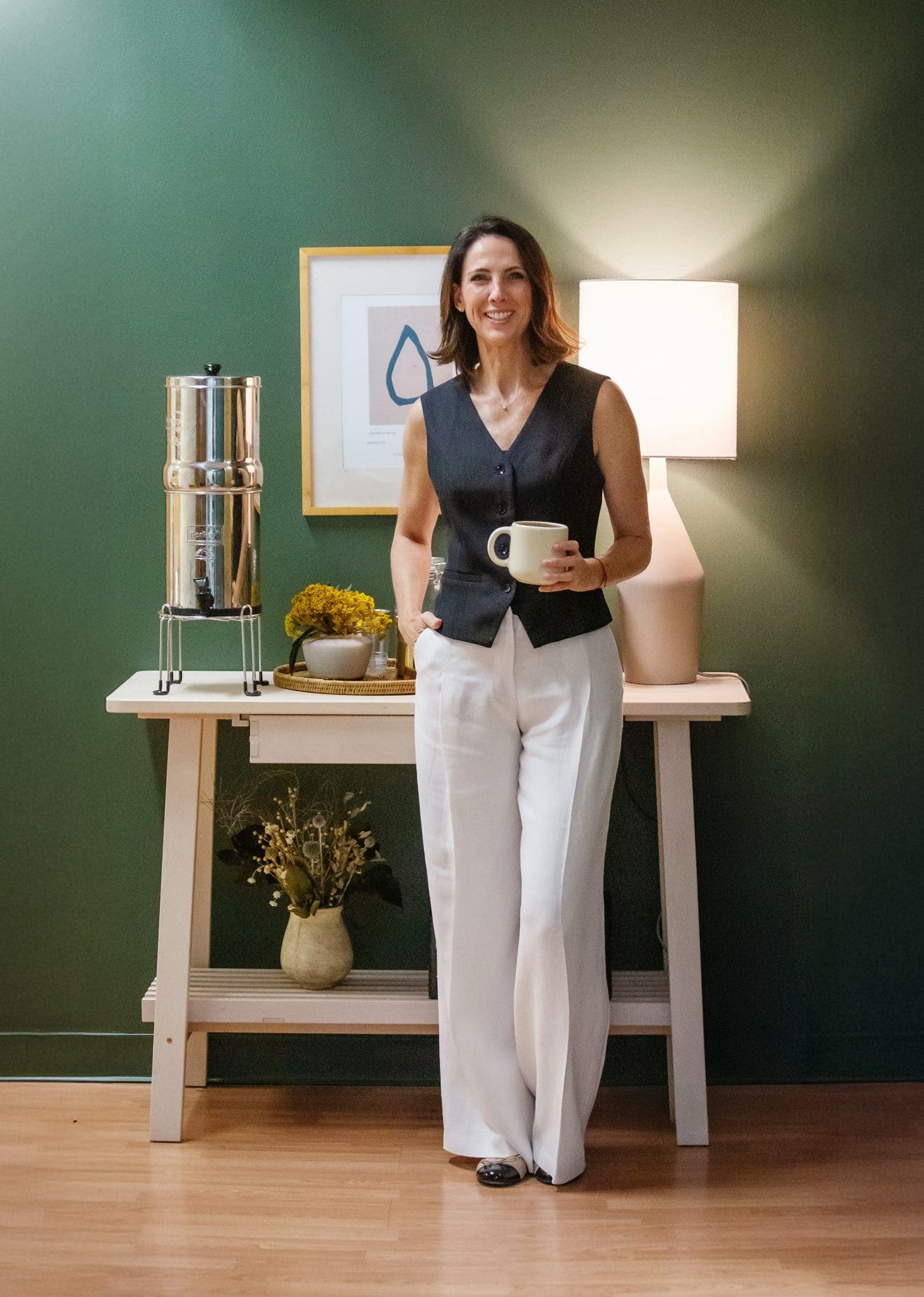 Woman holding mug, standing by a table with lamp.
